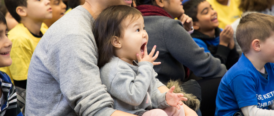 A little girl sits on her father's lap, excitedly watching a puppet show.