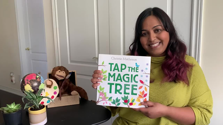 A smiling librarian sits next to a table holding the book Tap the Magic Tree. There are small potted plants, a globe and a stuffed toy monkey on the table.