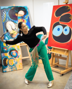 Blessyl Buan holds a paintbrush with several paintings in the background.